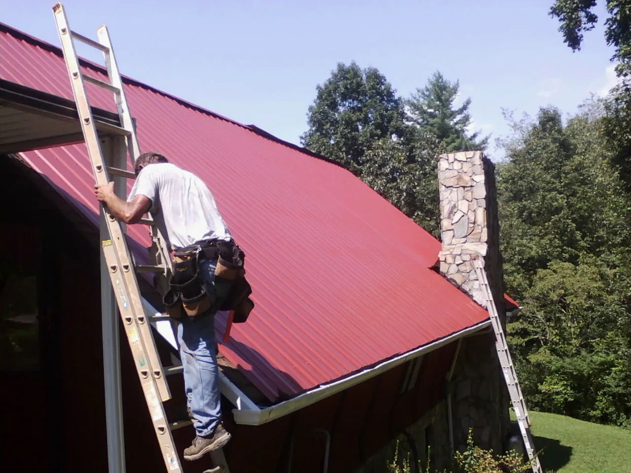 Roofer on a ladder beside a red metal roof and stone chimney — Soffit & Fascia Repair by Roofing Brothers in North Aurora